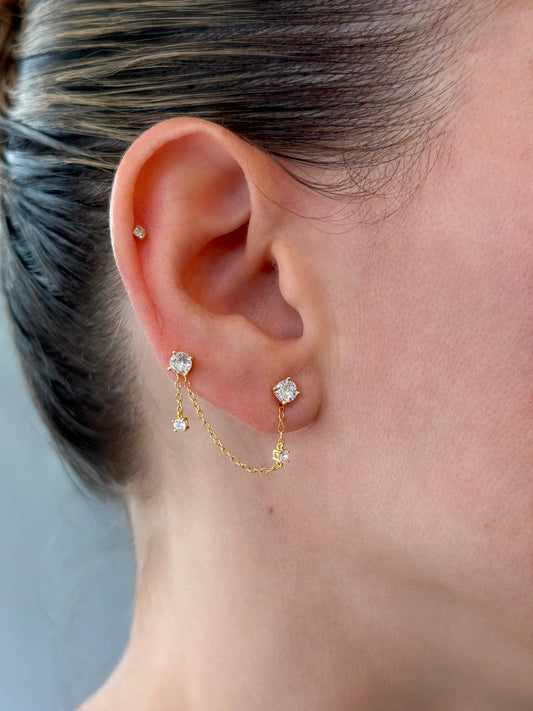 Sterling silver double piercing stud earrings connected by a dainty chain with a dangling charm on a model's ear