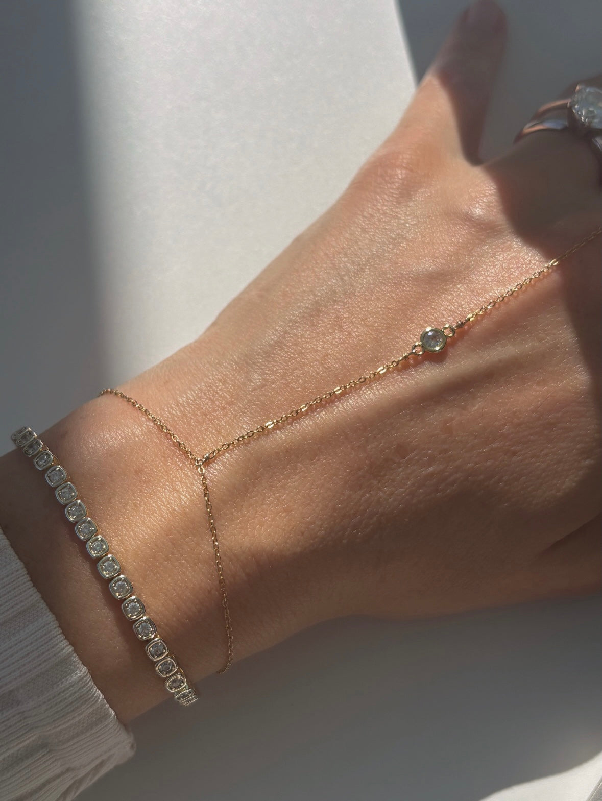 model hand wearing a dainty hand chain with bezel detail and a square tennis bracelet.