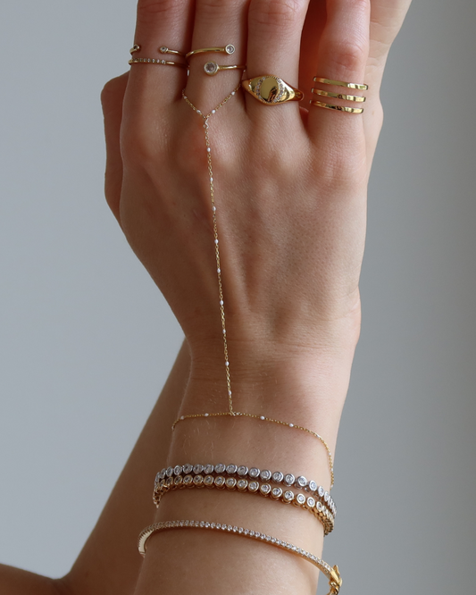 Model hand wearing a beautiful gold ring stack, a hand chain with white accents, 2 tennis bracelets and a thin pave bangle